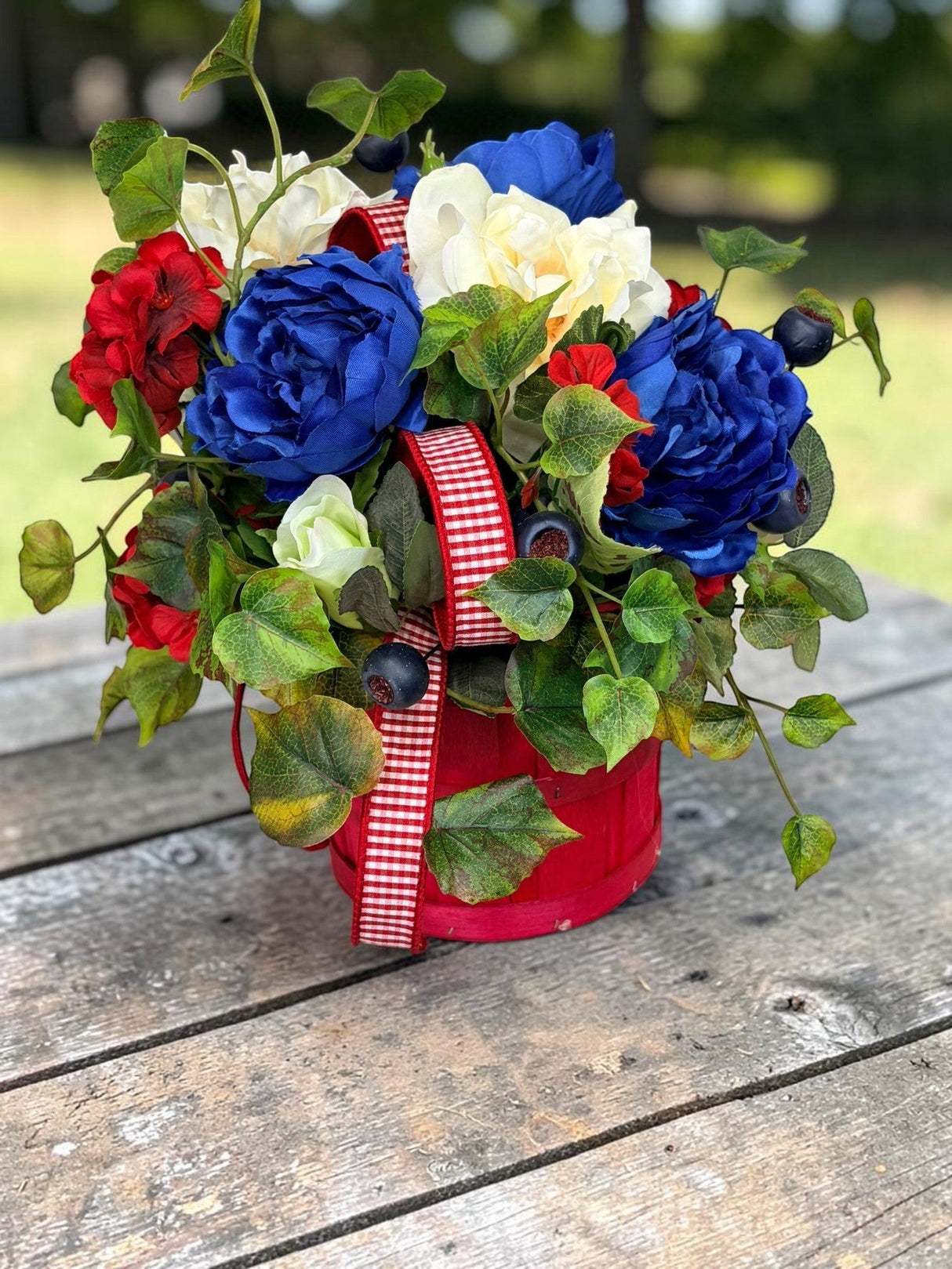 TCTCrafts patriotic floral arrangement in a red basket with red, white, and blue artificial flowers, greenery, and ribbon.
