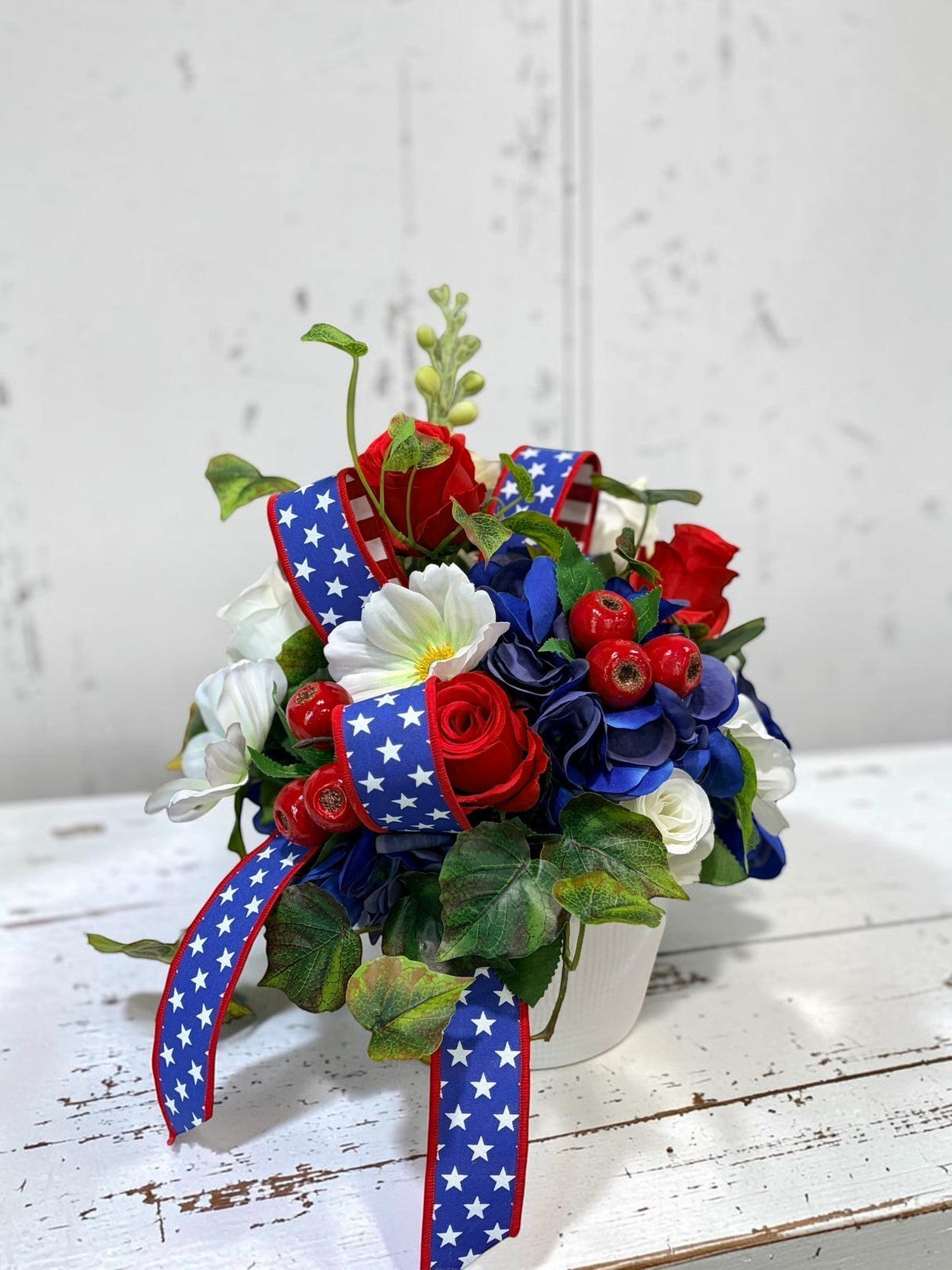 TCTCrafts patriotic floral arrangement with red roses, white cosmos, blue hydrangeas, and red berries in a white container with star-spangled ribbon.