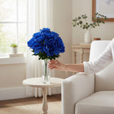TCTCrafts artificial blue peony bush with 12 blooms and green foliage in a clear vase on a side table.