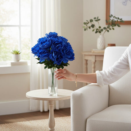 TCTCrafts artificial blue peony bush with 12 blooms and green foliage in a clear vase on a side table.