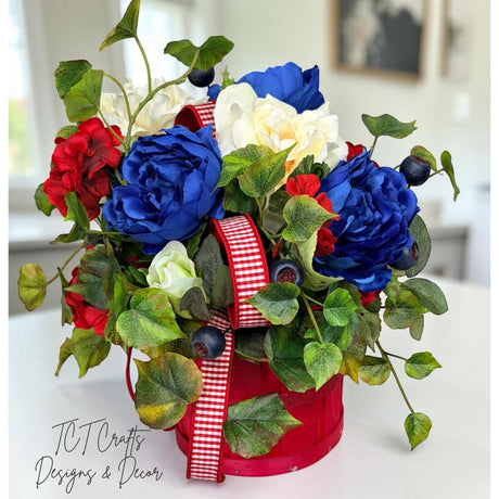 TCTCrafts patriotic floral arrangement in a red basket with red, white, and blue artificial flowers, greenery, and ribbon.