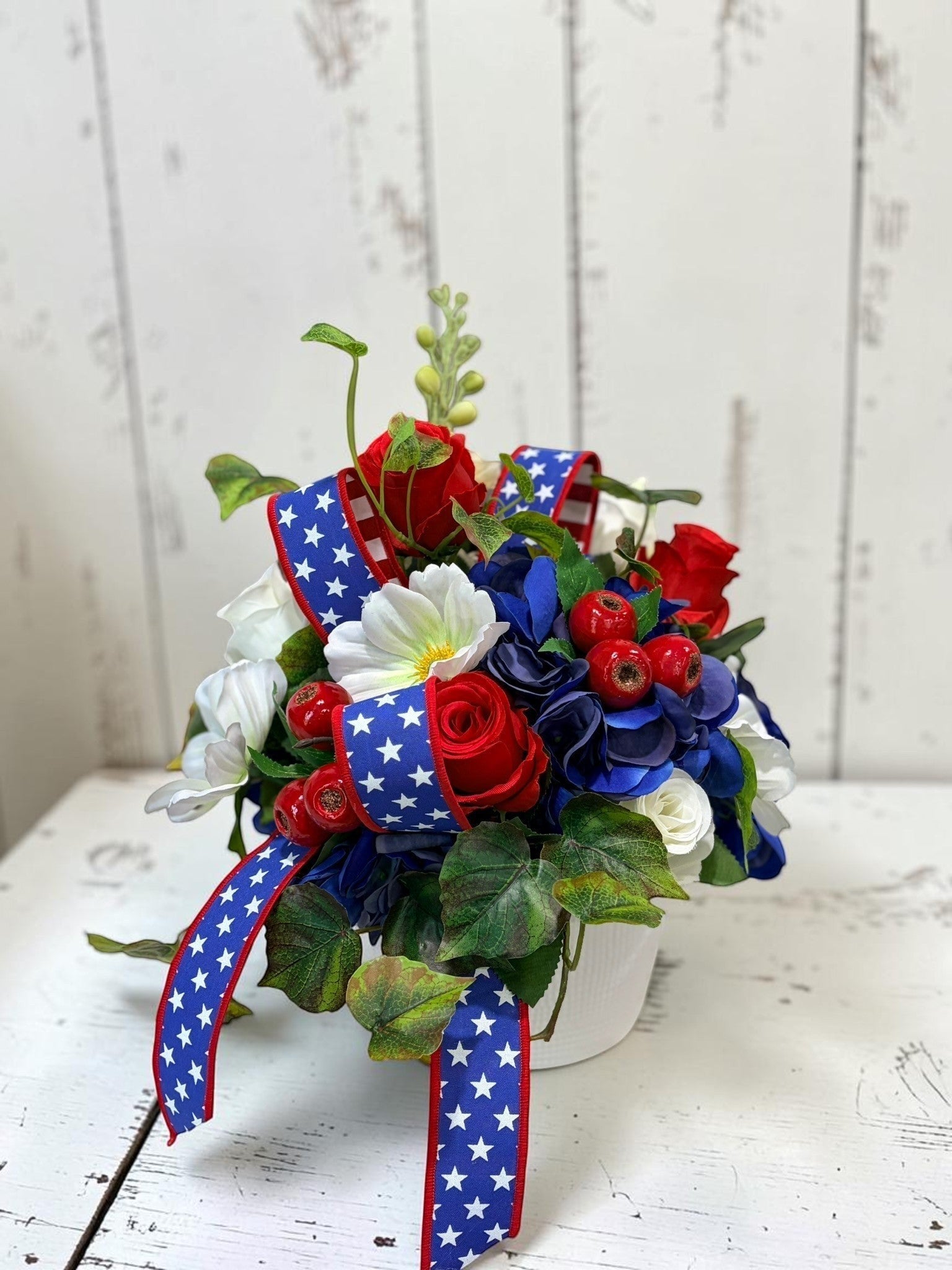 TCTCrafts patriotic floral arrangement with red roses, blue hydrangeas, white cosmos, red berries, and greenery in a white container with star-spangled ribbon.