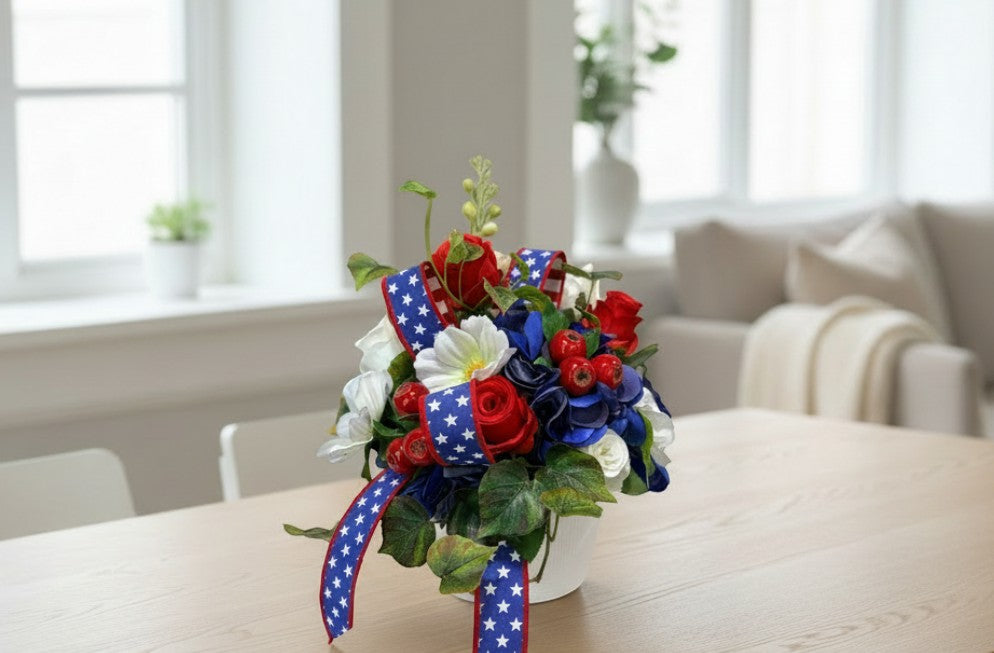 TCTCrafts patriotic floral arrangement with red roses, blue hydrangea, white cosmos, and greenery in a white container with star-spangled ribbon.