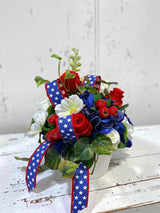 TCTCrafts patriotic floral arrangement with red roses, white cosmos, blue hydrangeas, and red berries in a white container with star-spangled ribbon.