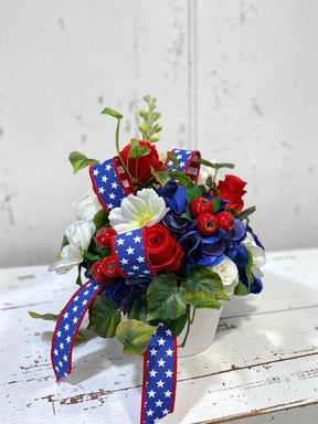 TCTCrafts patriotic floral arrangement with red roses, white cosmos, blue hydrangeas, and red berries in a white container with star-spangled ribbon.