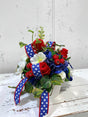TCTCrafts patriotic floral arrangement with red roses, white cosmos, blue hydrangeas, and red berries in a white container with star-spangled ribbon.